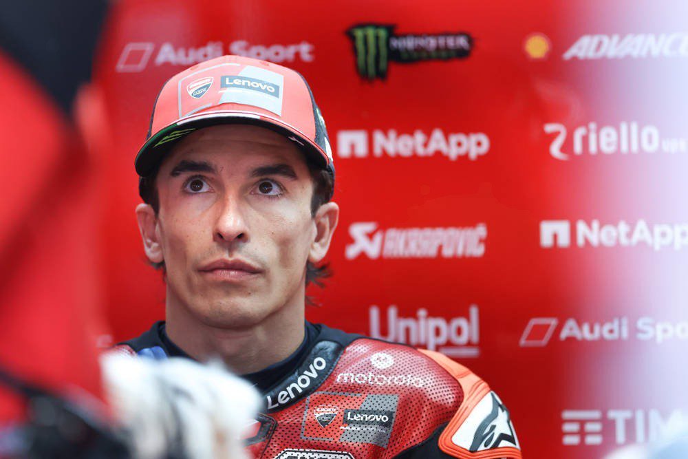 A rider in a red cap and red Ducati leathers, with Lenovo branding, in front of a red backdrop of sponsor logos in the MotoGP paddock