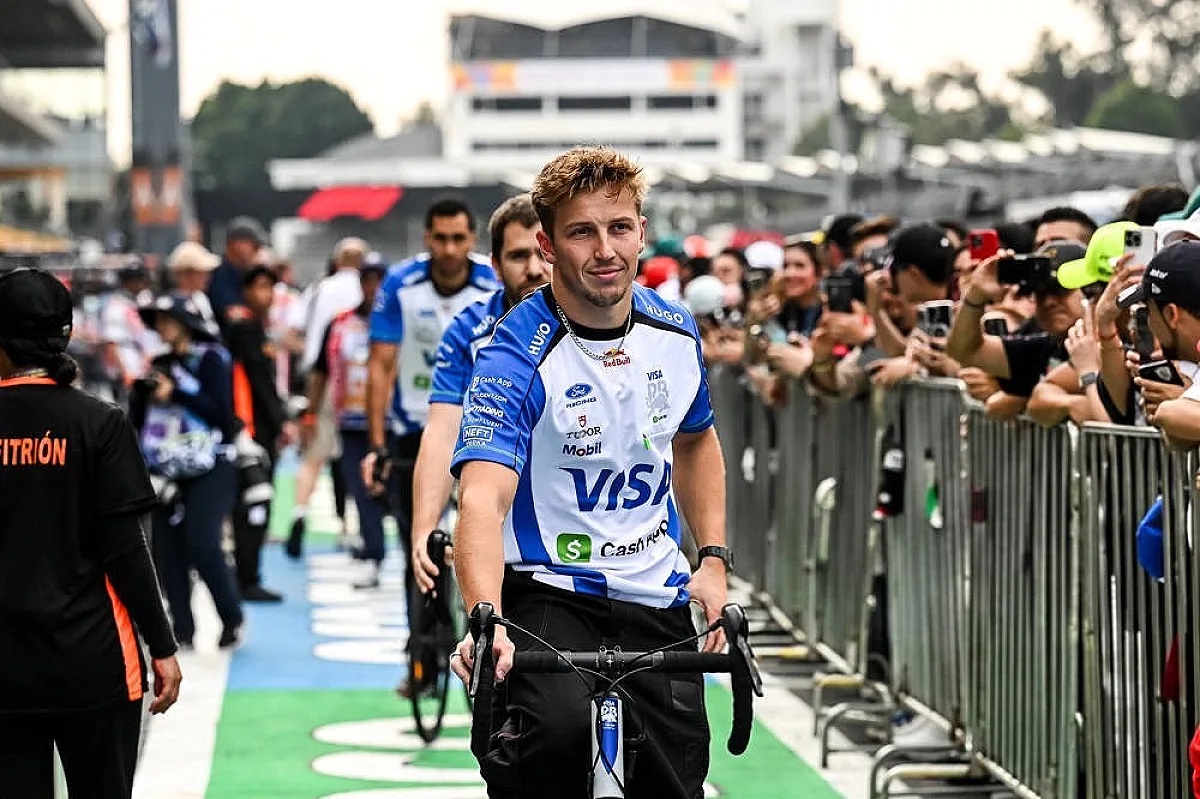 A Racing Bulls driver in blue and white team kit rides a bicycle through a Formula 1 paddock lined with fans and photographers