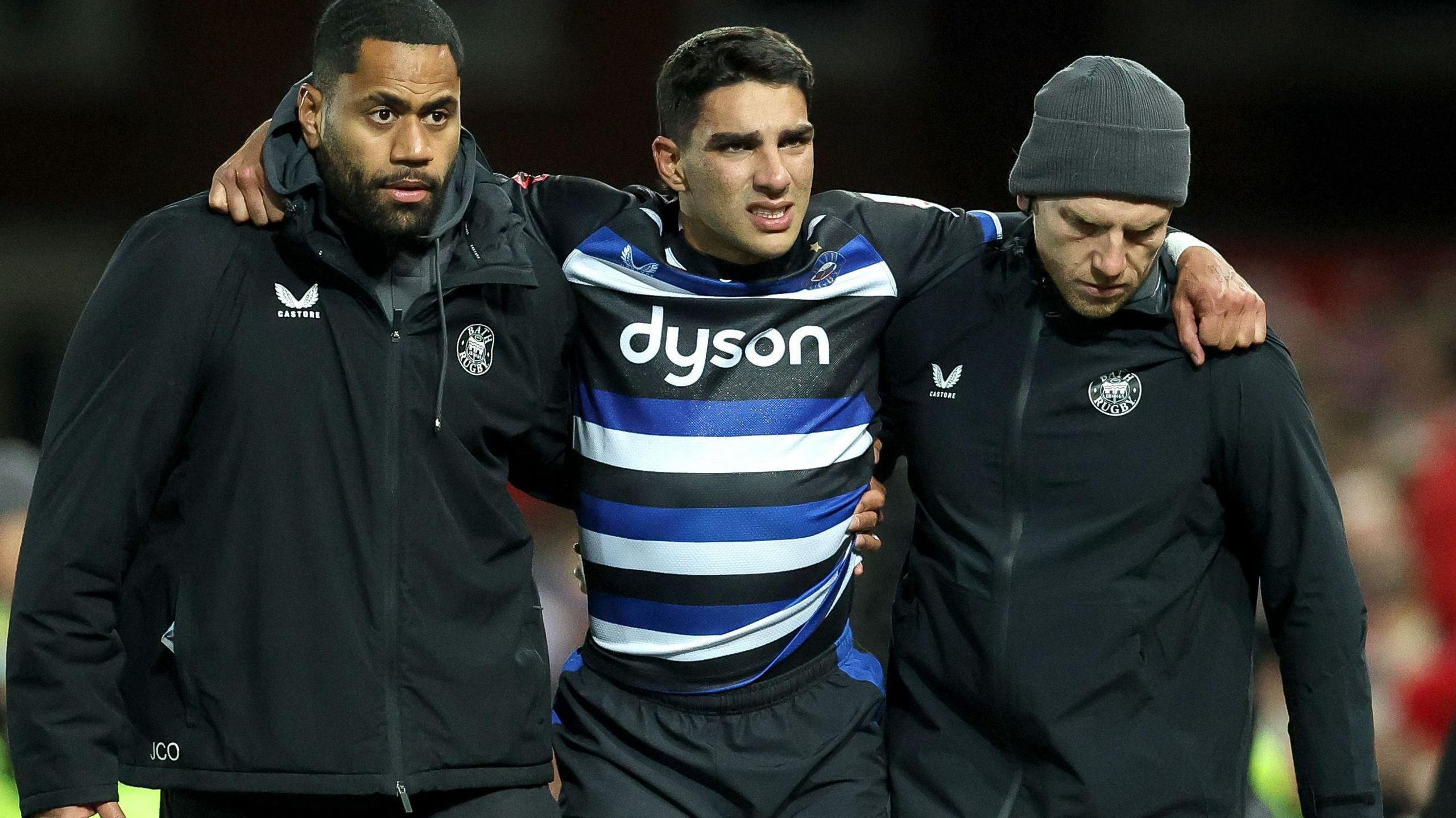 Santi Carreras in blue, white and black Bath shirt with arms round support staff as he is helped off the field