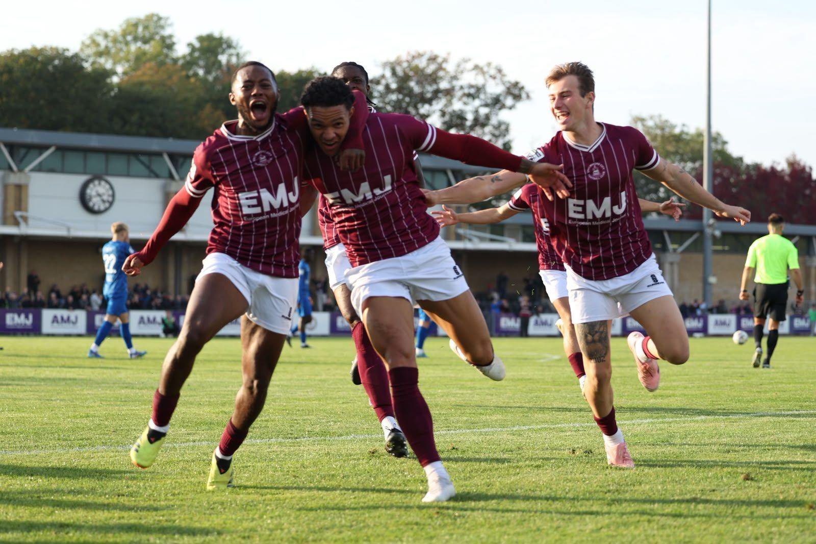 Male footballers dressed in claret shirts with white stripes are running towards the camera on a green football pitch. There is a stadium stand in the background.