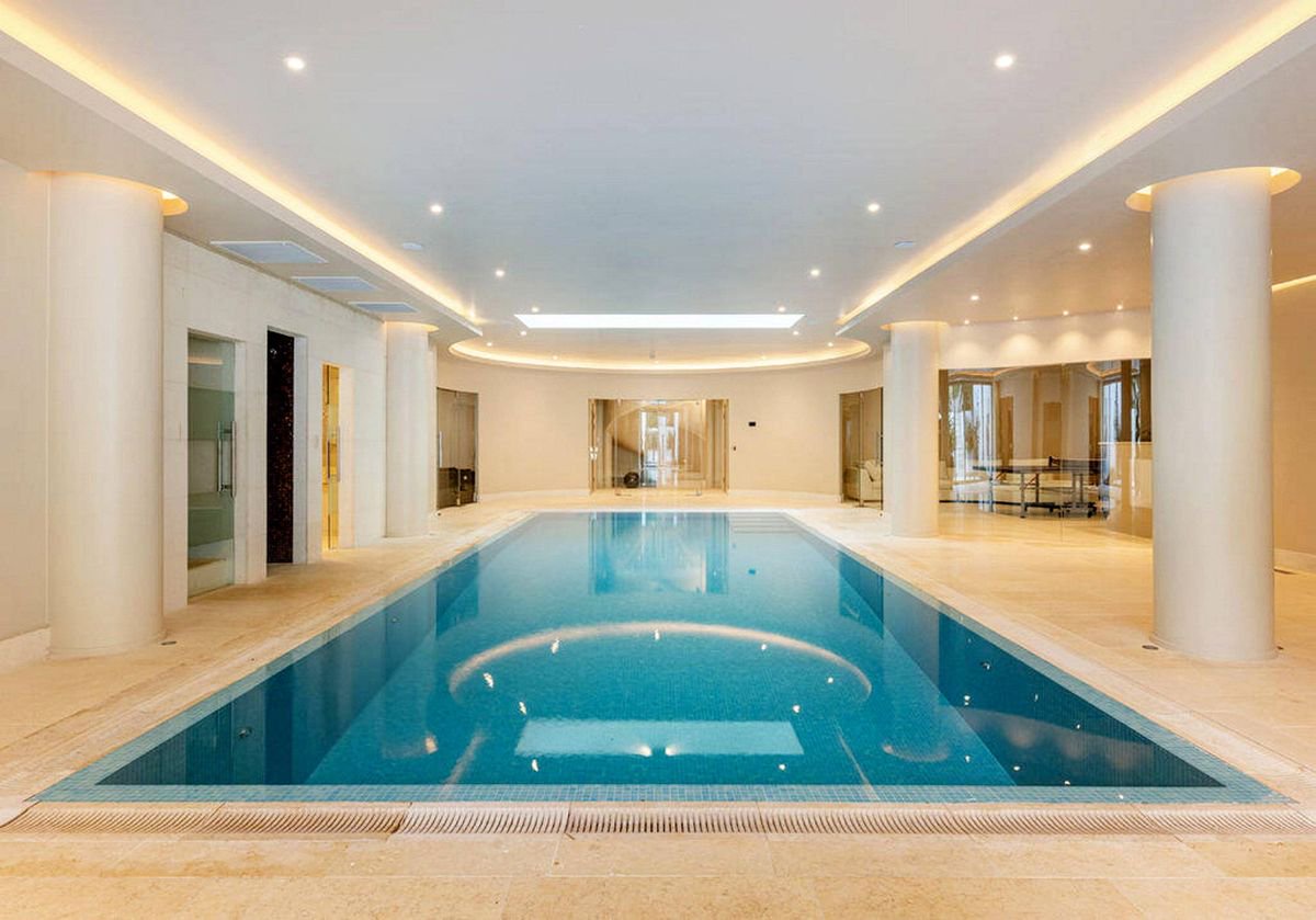 An indoor swimming pool with crystal clear blue water, surrounded by elegant white pillars, and adorned with modern lighting fixtures, creating a serene and sophisticated ambiance.