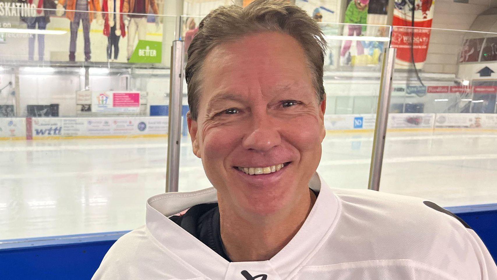 Image of a man wearing a white top, sitting on the edge of an ice rink and smiling at the camera