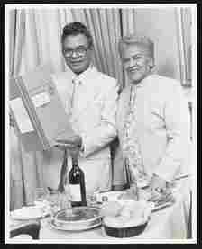 Black and white photo of a couple, dressed in 70s era business attire in front of a table of food at a restaurant. The man is holding up a menu.