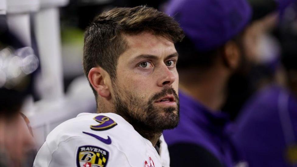Head and shoulders of Justin Tucker, wearing a white top, short dark hair, and dark beard