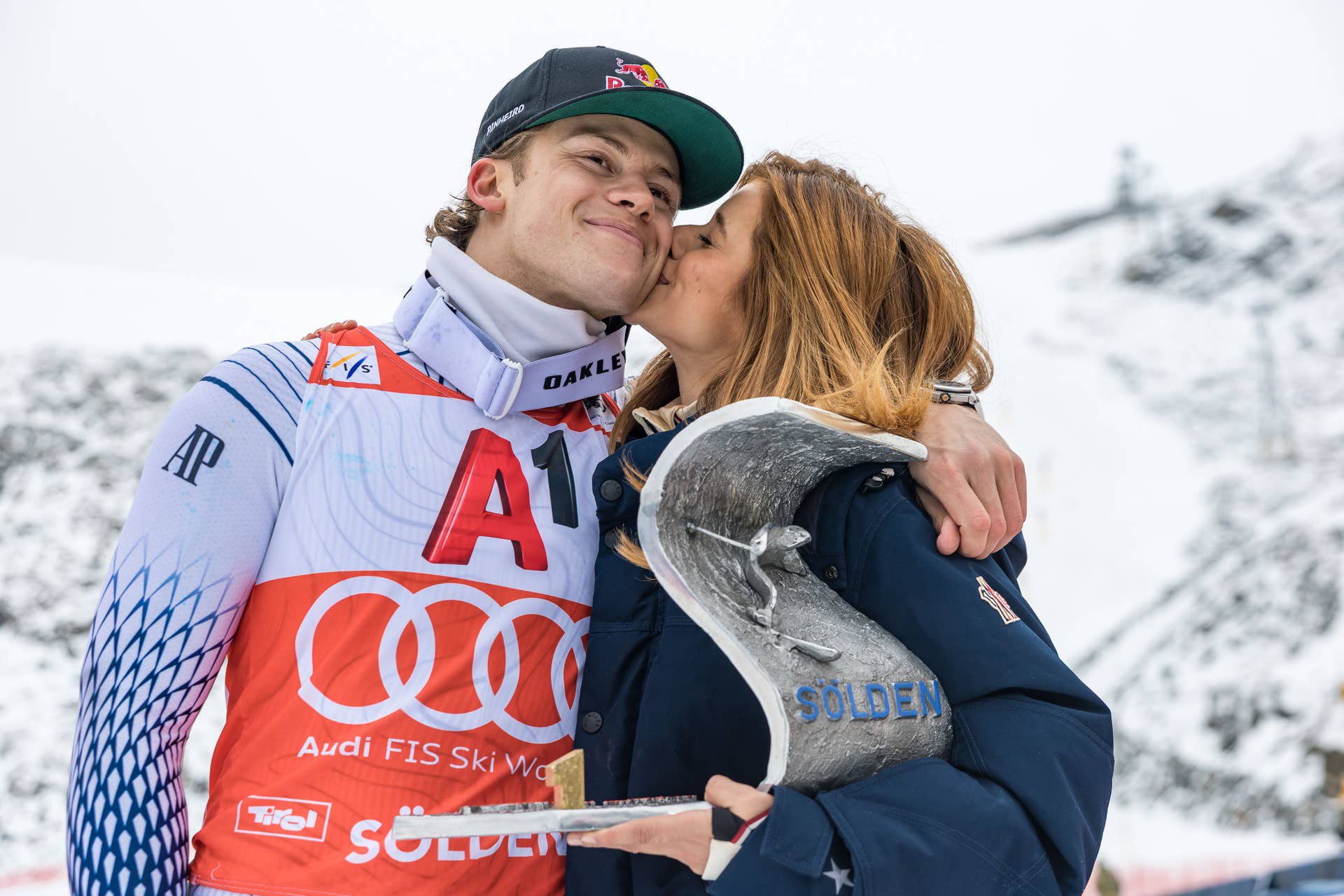 Ein Skifahrer wird bei der Siegerehrung von einer Frau auf die Wange geküsst, mit Pokal in der Hand.