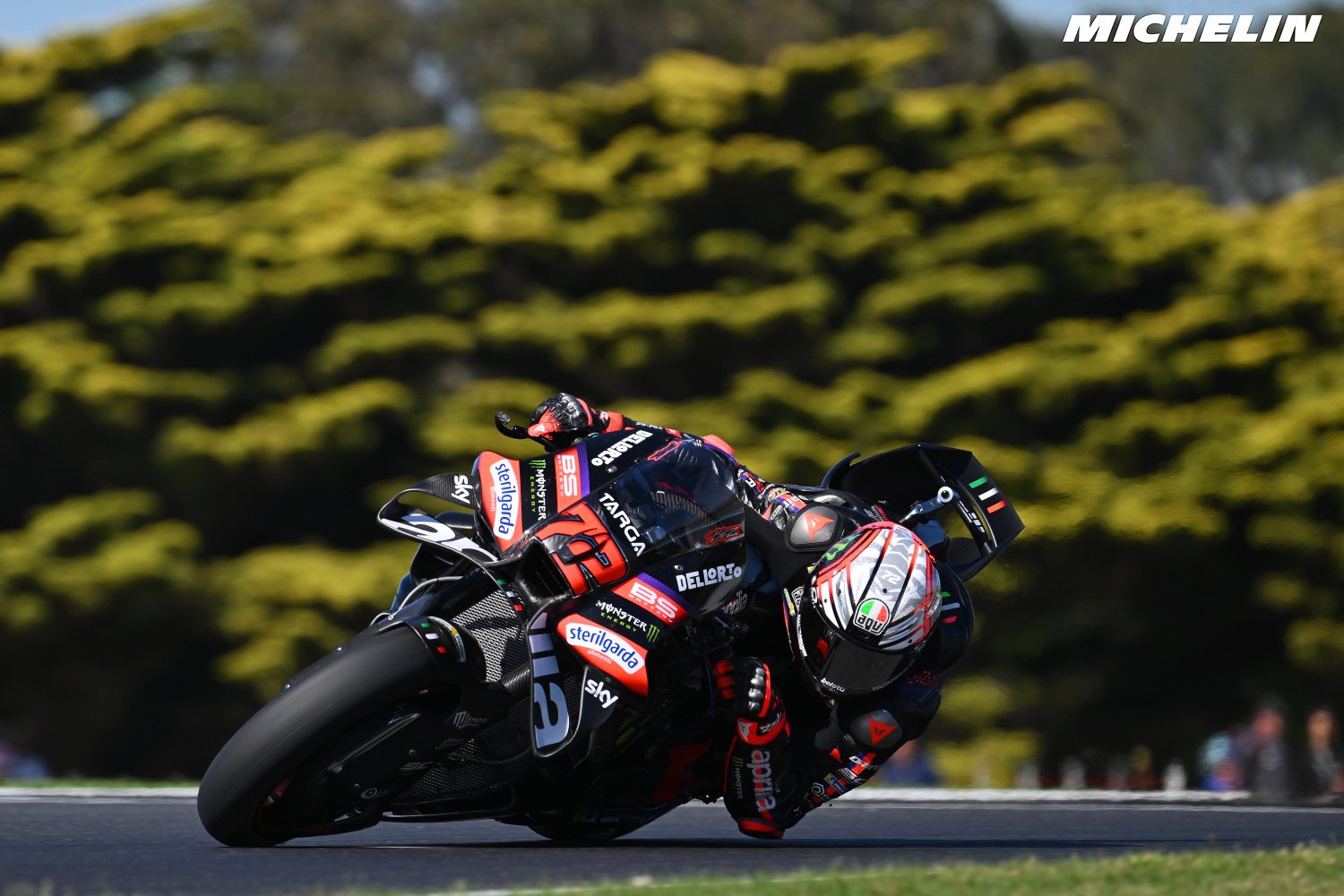 Rider in black and red Aprilia leathers on a MotoGP bike leaning into a corner, with trees and Michelin branding behind