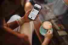 A person wearing a smartwatch holds a smartphone displaying financial management apps in one hand and a cappuccino in a saucer in the other hand, sitting at a desk