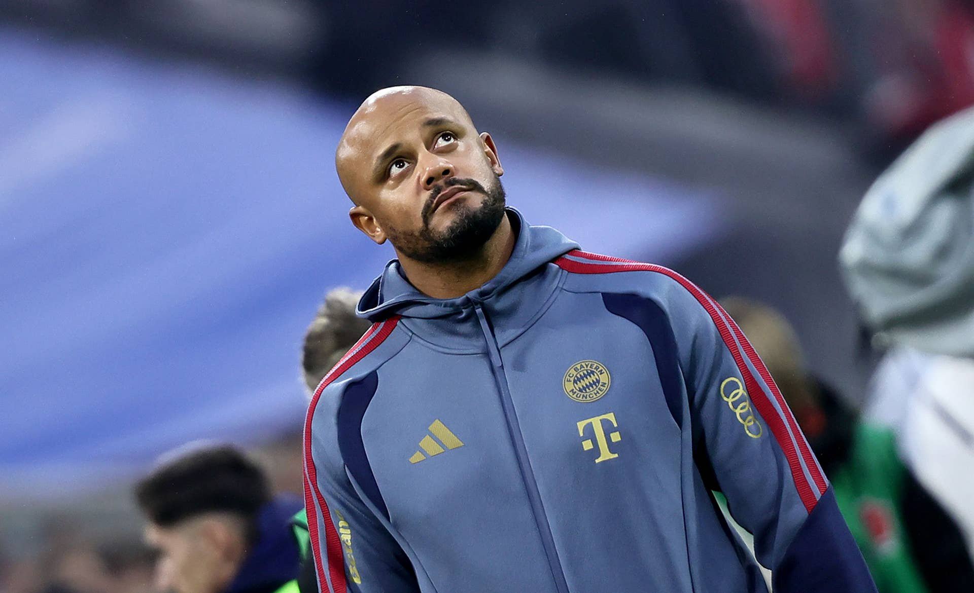 Ein Fußballtrainer im Bayern-München-Trainingsanzug blickt nachdenklich nach oben.