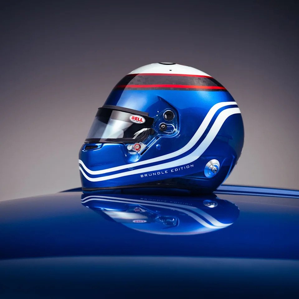 Blue racing helmet with white stripes and a glossy finish, reflecting on a surface, labeled "Brundle Edition.