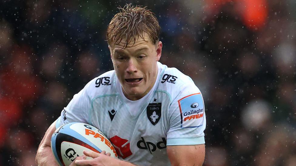 Fin Smith in a white Northampton Saints jersey during a match. He is carrying a rugby ball as rain falls around him.