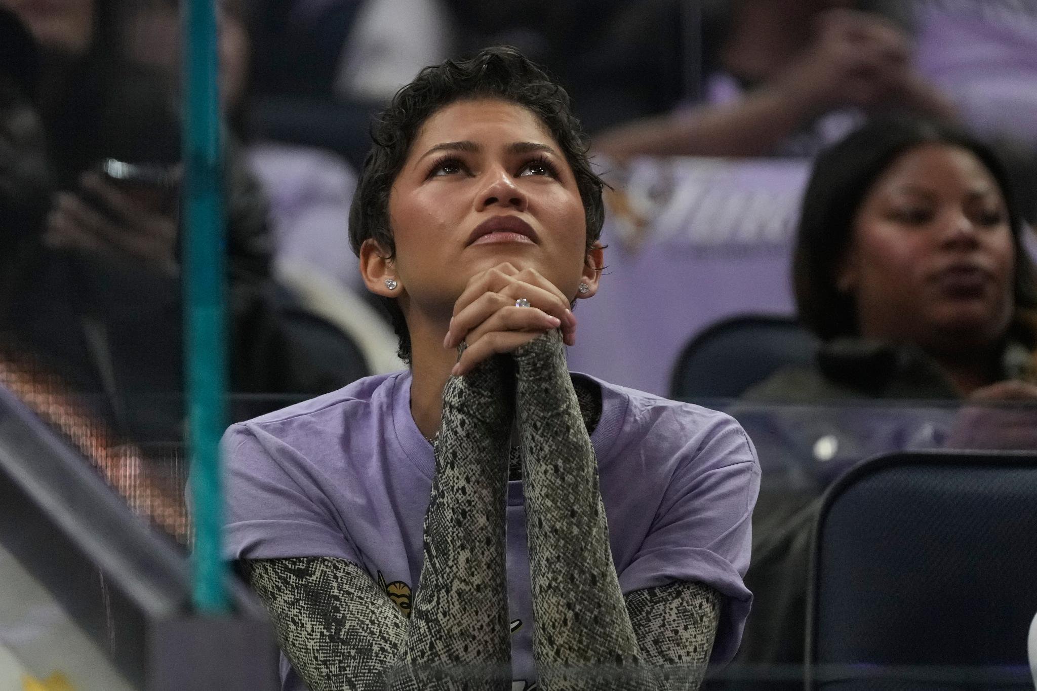 Zendaya watches intently with her hands clasped together during a WNBA basketball game.