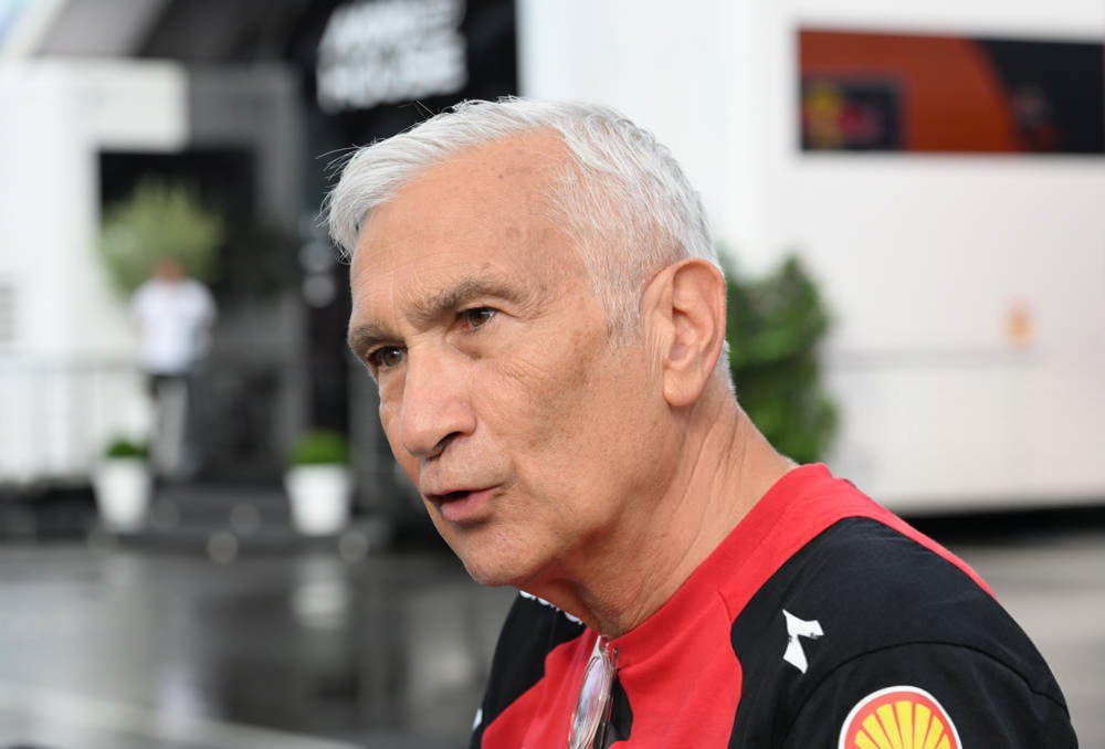 A grey-haired man in a red and black Shell-branded shirt speaks outdoors in a paddock area, with a team truck behind him.