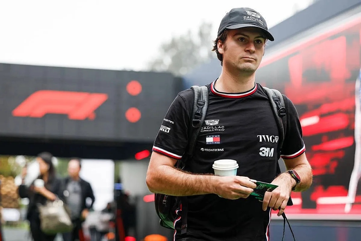 A man in a black Cadillac Formula 1 team cap and shirt, carrying a backpack and a coffee cup, walks through a paddock near a large F1 logo.