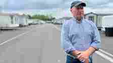 BBC Billy Welch wears a baseball cap, stands with clasped hands with a row of caravans behind him