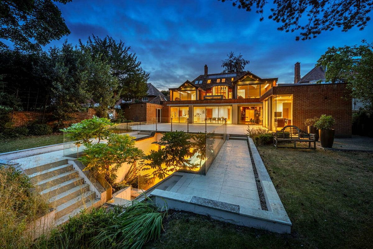 An elegant modern residence is highlighted at night, with its exterior lighting illuminating the well-manicured lawn and the sophisticated design of the house. A serene pond reflects the lights, creating a tranquil ambiance, surrounded by lush greenery.