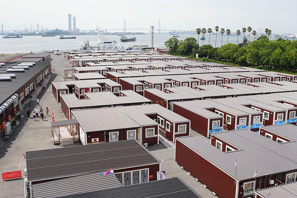 Aerial view of a large complex made up of rows of brown container-unit accommodation with grey roofs, arranged in a grid beside the waterfront in Nagoya. Several ships are visible in the harbour beyond the site, with a bridge, trees and palm trees in the background. A few people walk along the pathways between the buildings on a clear day.