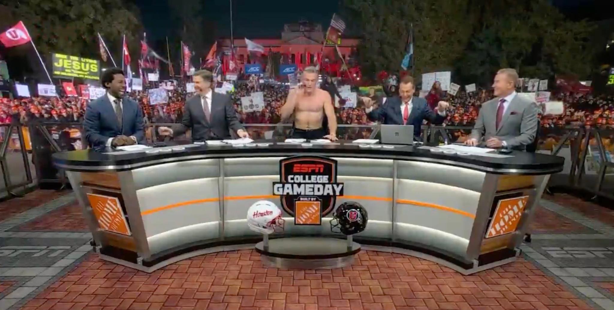 Pat McAfee stands shirtless behind the "College GameDay" desk alongside his fellow analysts in front of a large crowd.