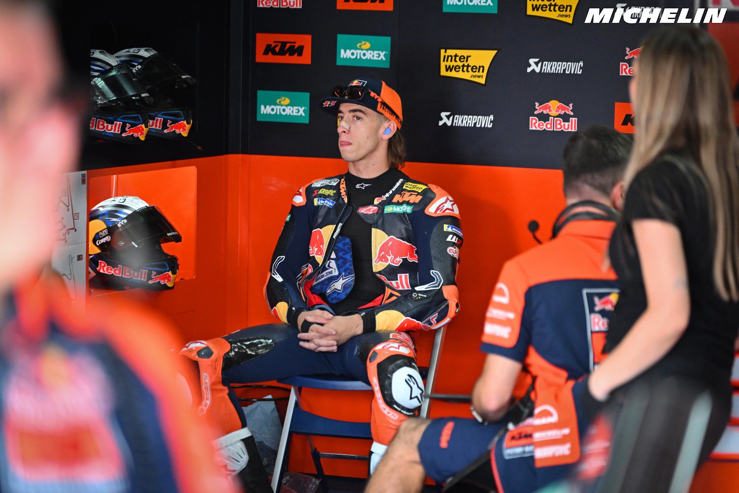 Red Bull KTM rider in race leathers seated in the team garage, with helmets and sponsor logos behind him (MotoGP)
