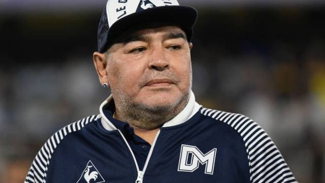 Diego Maradona, com barba grisalha, usando boné branco e azul marinho com a inscrição LE COQ SPORTIF e uma jaqueta esportiva azul marinho com listras brancas nos ombros e o logo DM no peito, olha para a direita com expressão séria