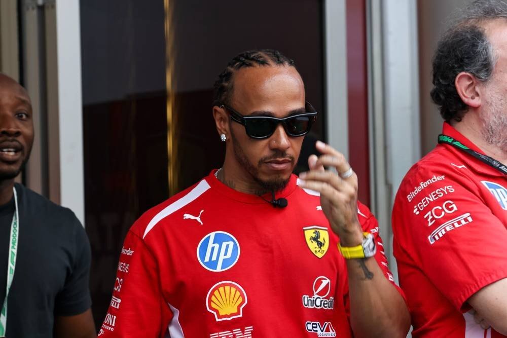 A man in sunglasses and a red Ferrari team shirt gestures while speaking in the F1 paddock, with a Shell and HP logo visible.