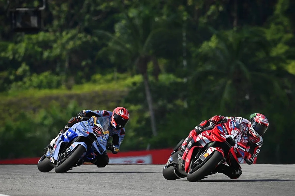 Two MotoGP riders on a track: one in red and white Ducati livery, another on a blue Yamaha-style bike, cornering past lush green palms at Malaysia's circuit.