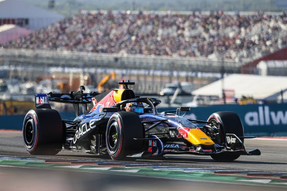 Red Bull F1 car with a driver in a cockpit on a track at Austin, with crowded grandstands behind and "Oracle" sponsorship on the side.