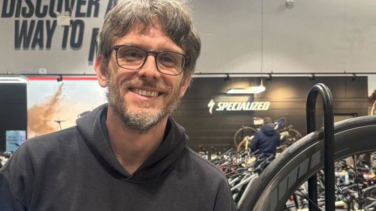 Matthew Porter has black and grey hair and beard and is wearing dark framed glasses and a black hooded top. He is stood inside a shop with rows of bikes behind hi,