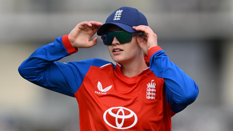 England Women's stand-in captain Charlie Dean stands in the outfield during a game, wearing a blue England cap and black sunglasses