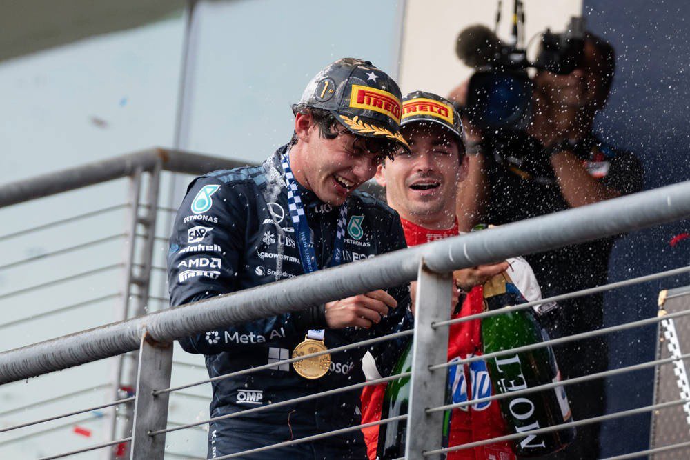 Two drivers on a podium spraying champagne; one wears a Mercedes-AMG Petronas team jacket and gold medal, the other a red Ferrari-style suit, at a Formula 1…