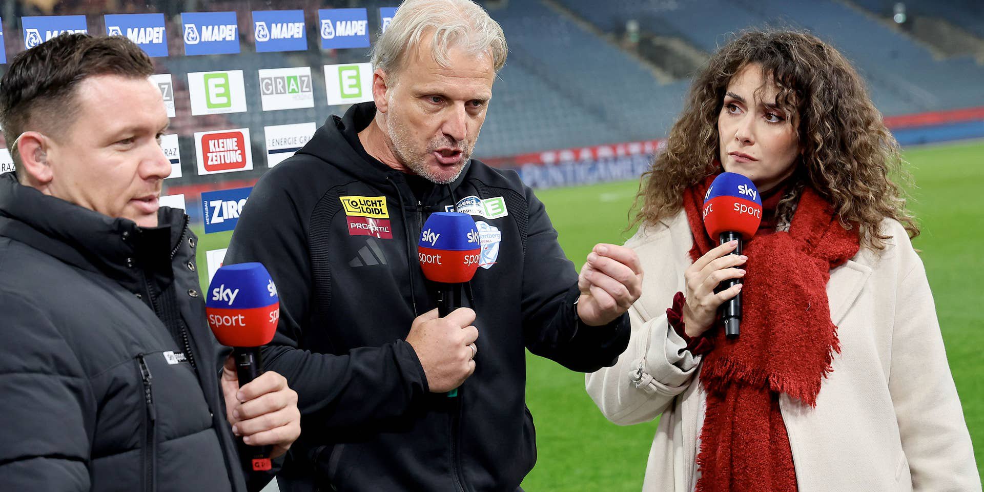 Drei Personen mit Sky-Mikrofonen führen ein Interview im Fußballstadion.