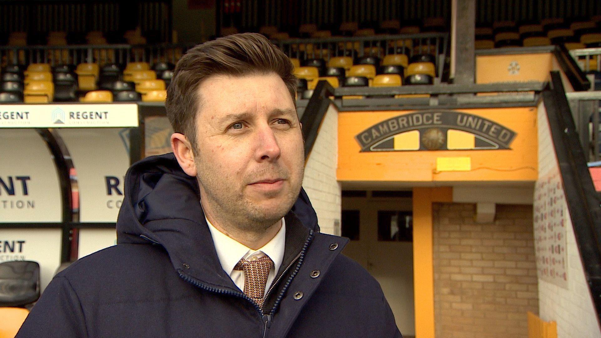 Alex Tunbridge standing in front of the tunnel at the Abbey Stadium. He is wearing a blue jacket, which is zipped up, a white shirt and brown tie. The pitchside tunnel behind is painted yellow with the club's crest above.