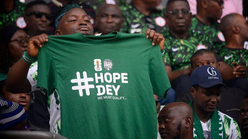 A Nigeria football fan holds a green T-shirt with the words 'Hope Dey' (meaning 'there is hope') written in white and 'we must qualify' underneath. He is surrounded by other Nigeria fans who are seated in a stadium 