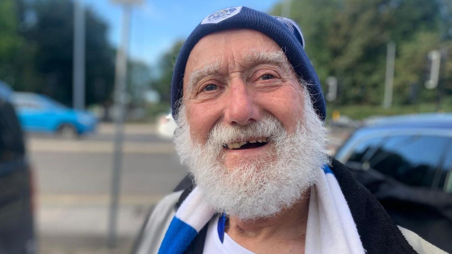 A close-up portrait of Mike Hamilton wearing a blue-and-white scarf and a navy hat, standing beside a roadside with trees and passing traffic blurred in the background. He has a white beard.