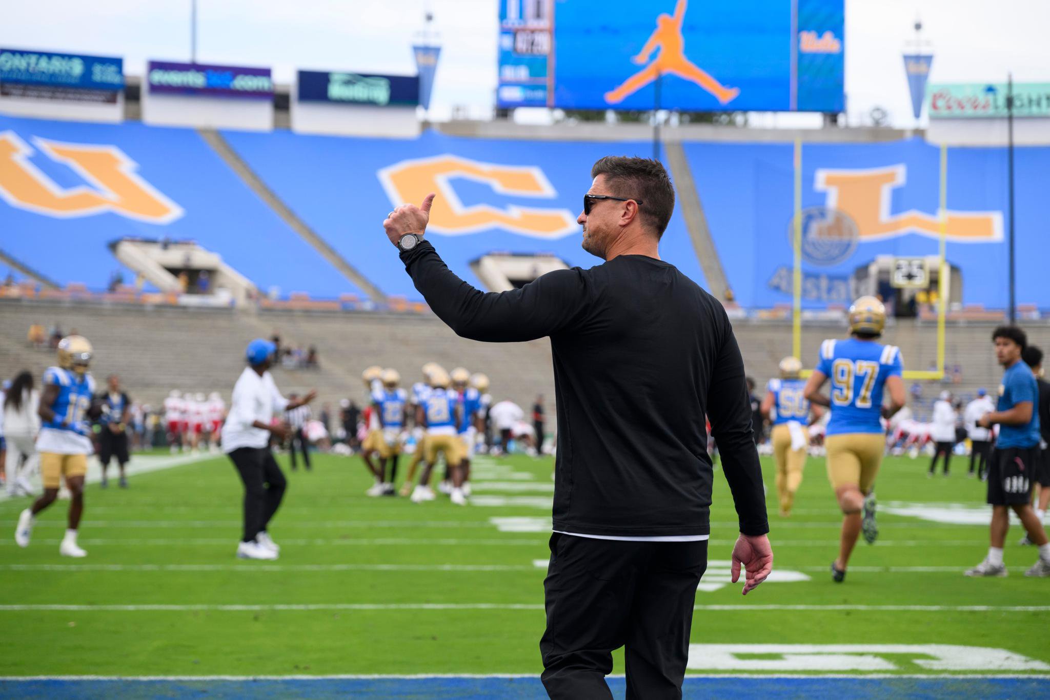 Football coach gesturing on the field during practice at a stadium.
