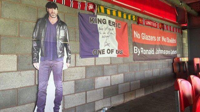 A photo showing a cardboard cut-out of former Manchester United player Eric Cantona, next to a French flag with the words 'King Eric one of us' written on it, leaning against a wall at the back of a stand at Broadhurst Park.
