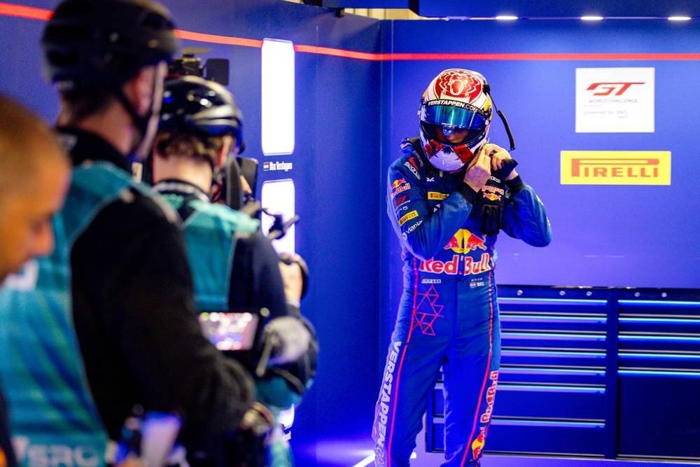 Driver in blue Red Bull racing suit and helmet adjusting his gear in a garage, with camera crews filming nearby