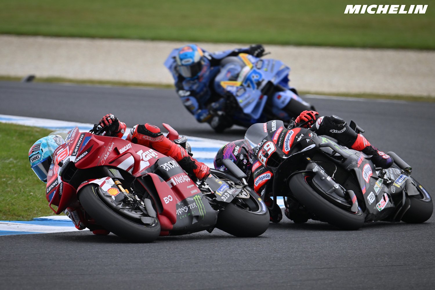 Ducati rider in red leathers and Ducati Lenovo-style livery leaning into a corner ahead of a rival Aprilia in black, MotoGP race, Michelin branding visible.