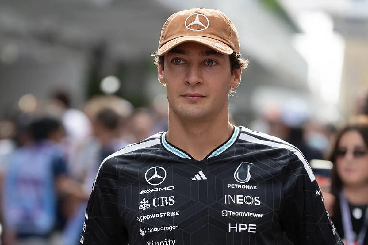 Driver in a black Mercedes-AMG team shirt and tan cap in a paddock setting, Formula 1 team kit with Petronas and INEOS sponsor logos.