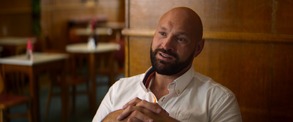 Tyson Fury sitting in an indoor setting with wooden paneling, speaking in a candid interview.