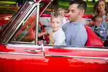 A man drives a vintage car with a young boy in his lap. Another woman and two children are in the backseats