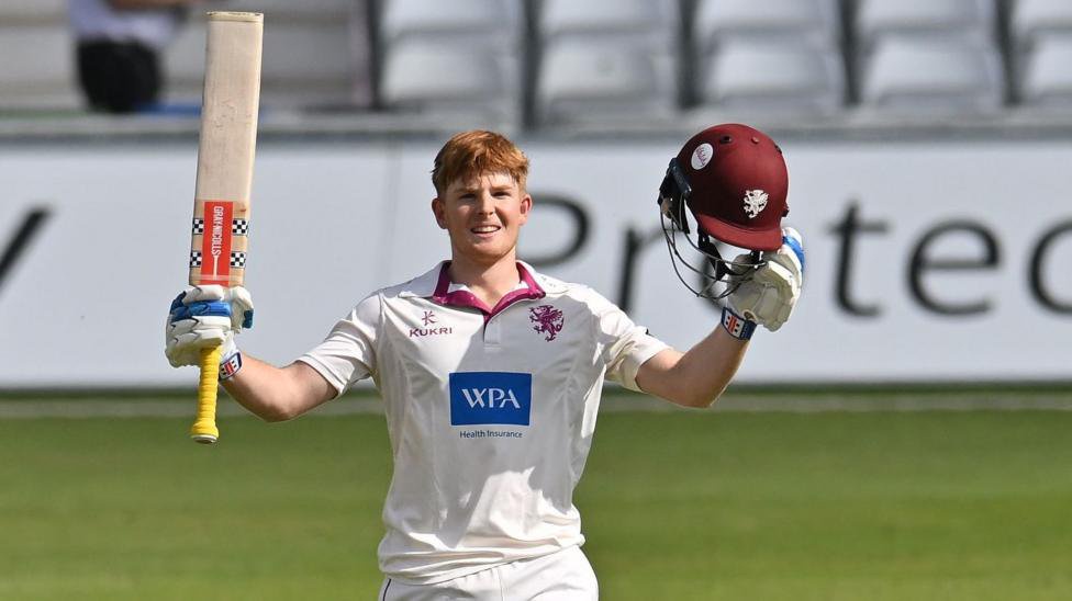 Thomas Rew walks on the pitch holding his bat aloft in one hand and his helmet in the other. He is wearing white Somerset kit