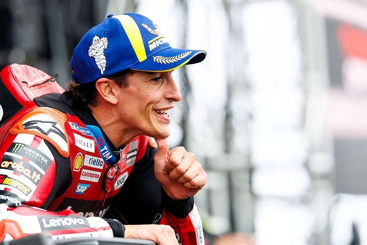 A Ducati MotoGP rider in red and black leathers with Lenovo and Shell logos, smiling and giving a thumbs up in the paddock while wearing a blue Michelin cap.
