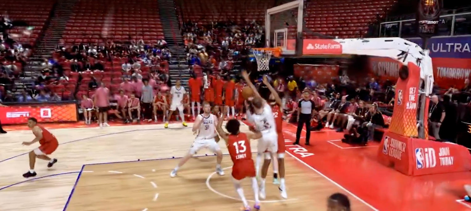 Basketball players compete under the net during a game in a packed arena with red seating.