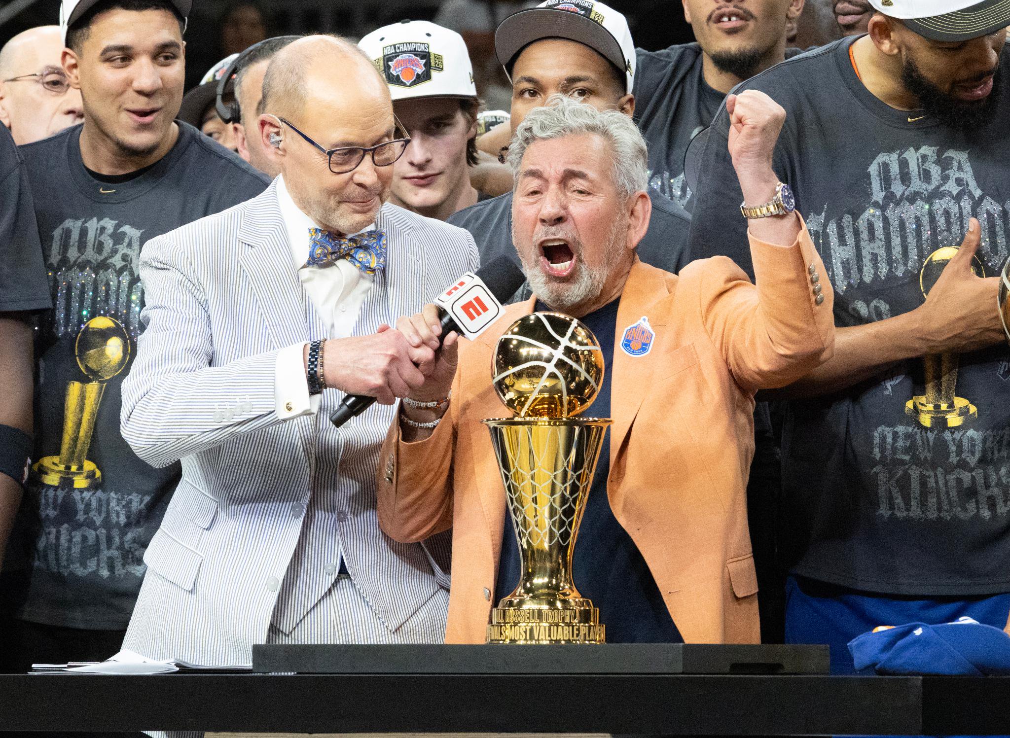 A man in an orange suit yells with joy, raising his fist next to the NBA championship trophy, while being interviewed with an ESPN microphone in front of New York Knicks players.