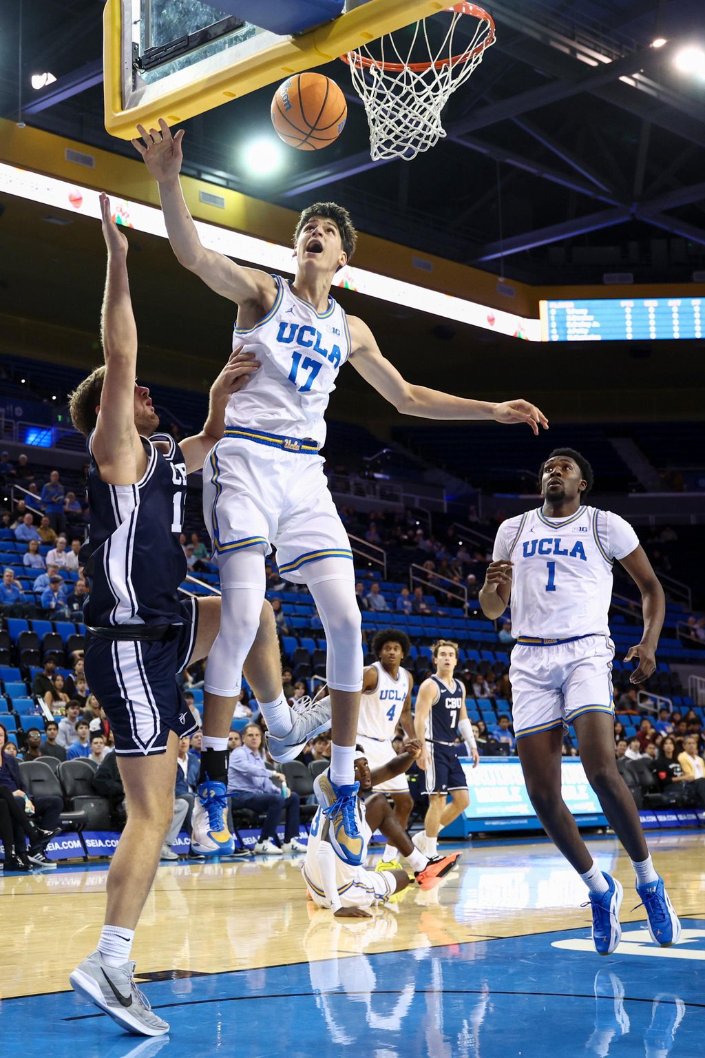 A UCLA basketball player in a white uniform leaps toward the basket while being defended by a Cal Baptist player in a dark blue uniform.