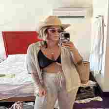 Woman in a crochet outfit with a bikini top underneath, wearing a straw hat and sunglasses, taking a mirror selfie in a messy room