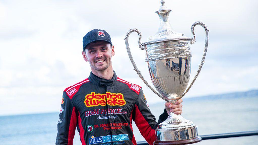 Osian Pryce holding the British Rally Championship trophy, in his black and red race suit and hat.