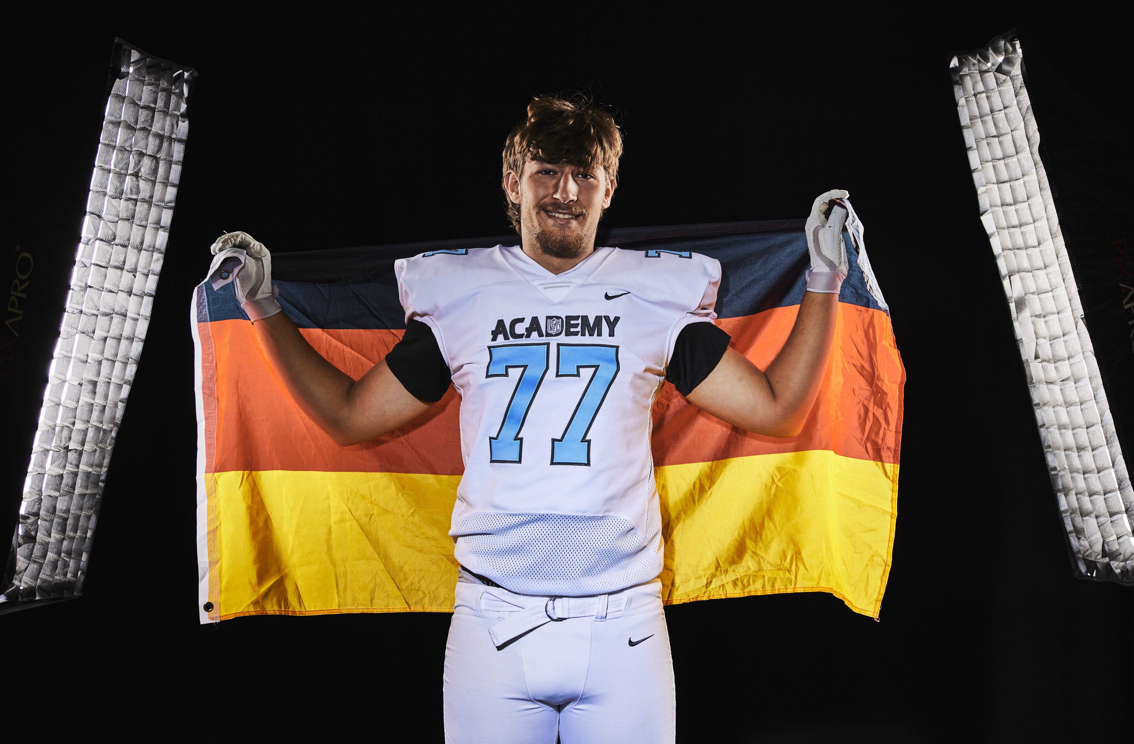 A football player in a white “ACADEMY 77” jersey holds a German flag behind him and stands between two vertical lights against a black background—offensive line prospect Niko Kampas proudly showcases his skills, representing both Tennessee and his heritage. Alt text generated by AI.