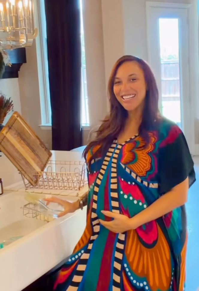 A pregnant woman wearing a colorful, butterfly-patterned kaftan stands in a kitchen, smiling and holding her belly while standing by the sink.