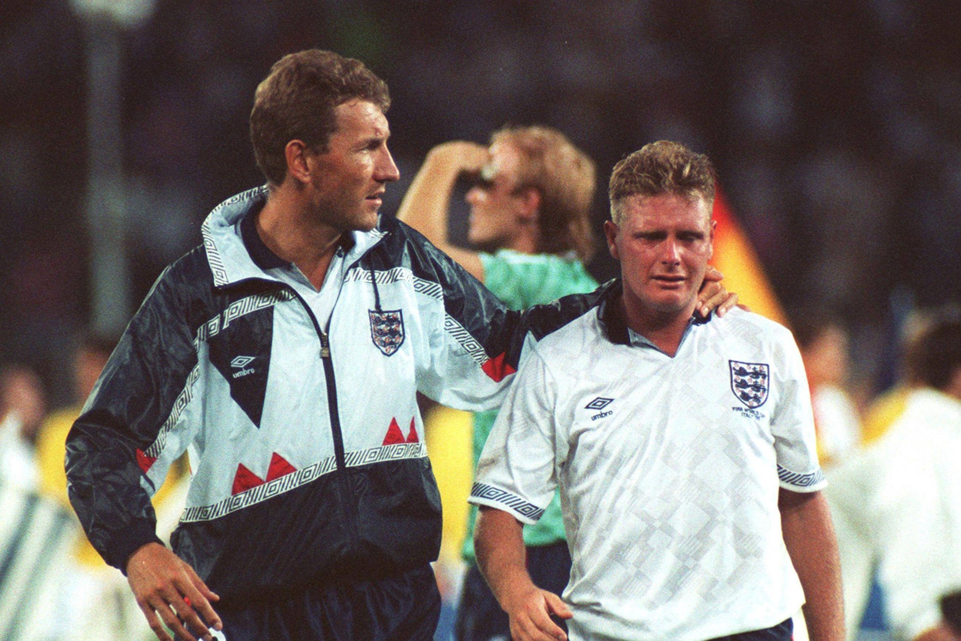 Terry Butcher in football kit and a football training jacket, walks on a football pitch, towards the camera, with his arm around the neck of Paul Gascoigne, who is on his left. Gascoigne wears a white England shirt, with the Three Lions' crest, and he is crying.